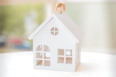 Beyaz minyatür ev modeli parlak, bulanık bir arka planı olan bir masada oturuyor. Bu sembolik imaj gayrimenkul, ev sahipliği, emlak yatırımı, ipotek ve ev sahibi olma hayalini temsil ediyor.