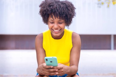 Kıvırcık siyah saçlı ve sarı bluzlu neşeli genç bir kadın açık havada oturmuş, akıllı telefonuna bakarken gülümsüyor. Ağaçların ve binaların bulanık şehir arkaplanı modern bir dokunuş katıyor..