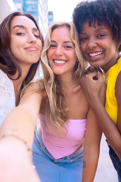 Çeşitli özelliklere sahip üç genç kadından oluşan bir grup, şehir ortamında selfie çekerken gülümsüyor. Sıradan yaz kıyafetleri giyip şehirde eğlenceli bir anın tadını çıkarıyorlar..
