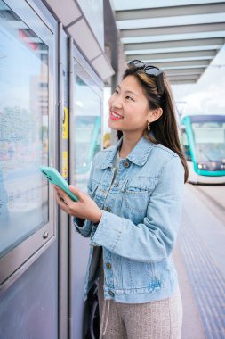 Kot ceketli, tramvay durağında bilet makinesi kullanan gülümseyen genç bir kadın. Dokunmatik ekranla iletişim kurarken elinde bir akıllı telefon tutuyor. Arka planda toplu taşımacılık bulunan kentsel ayar.