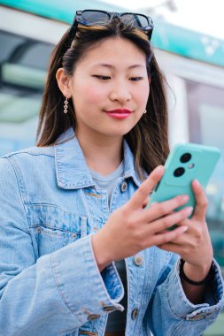  Tramvay durağında oturan kot ceketli neşeli genç kadın yaklaşan tramvayı beklerken akıllı telefonunu kullanıyor. Modern şehir ulaşımı ve dijital yaşam tarzı kavramı. Dikey