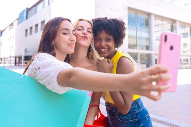 Çeşitli özelliklere sahip üç genç kadından oluşan bir grup, şehir ortamında selfie çekerken gülümsüyor. Sıradan yaz kıyafetleri giyip şehirde eğlenceli bir anın tadını çıkarıyorlar..