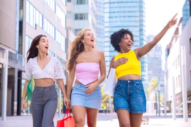 Üç genç kadından oluşan bir grup, alışveriş çantaları taşıyarak modern bir şehir caddesinde yürüyorlar. Bir tanesi heyecanlı bir şekilde bir şeye işaret ediyor hepsi gülümserken, eğlenceli ve hareketli bir alışveriş gününün tadını çıkarırken..
