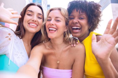 Çeşitli özelliklere sahip üç genç kadından oluşan bir grup, şehir ortamında selfie çekerken gülümsüyor. Sıradan yaz kıyafetleri giyip şehirde eğlenceli bir anın tadını çıkarıyorlar..