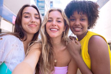 Çeşitli özelliklere sahip üç genç kadından oluşan bir grup, şehir ortamında selfie çekerken gülümsüyor. Sıradan yaz kıyafetleri giyip şehirde eğlenceli bir anın tadını çıkarıyorlar..