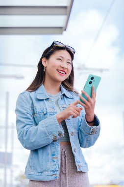 Kot ceketli neşeli genç bir kadın mutlu bir ifadeyle yukarı bakarken akıllı telefonunu tutuyor. Modern kentsel ortam, dijital bağlantıyı ve şehir yaşam tarzını temsil ediyor.