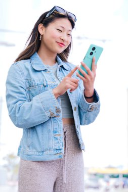 Kot ceketli neşeli genç bir kadın mutlu bir ifadeyle yukarı bakarken akıllı telefonunu tutuyor. Modern kentsel ortam, dijital bağlantıyı ve şehir yaşam tarzını temsil ediyor.