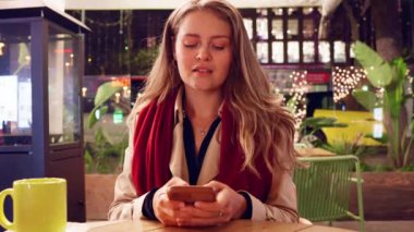  Genç bir kadın gece açık bir kafede oturuyor, akıllı telefonunu tutarken gülümsüyor. Sıcak atmosfer, şehir ışıkları ve modern kentsel ortam bir dijital bağlantı ve gevşeme anını vurguluyor.