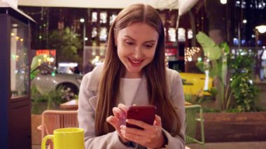  Genç bir kadın gece açık bir kafede oturuyor, akıllı telefonunu tutarken gülümsüyor. Sıcak atmosfer, şehir ışıkları ve modern kentsel ortam bir dijital bağlantı ve gevşeme anını vurguluyor.