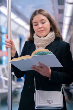 Modern bir metro treninde duran siyah paltolu ve bej eşarplı odaklanmış genç bir kadın metal bir direğe tutunarak kitap okuyor. Seyahat sırasında sakin bir anın tadını çıkarıyor..