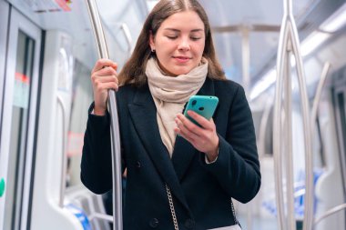 Siyah paltolu ve bej eşarplı neşeli genç bir kadın modern bir metro treninin içinde elinde akıllı bir telefon ve destek için metal bir direği tutuyor. Seyahat ederken ışıl ışıl gülümsüyor.. 