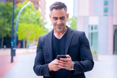 Mavi tişörtlü, gülümseyen orta yaşlı bir işadamı modern bir şehir ortamında akıllı telefon kullanıyor..