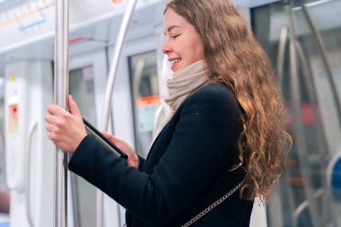  Modern bir metro treninde dururken siyah paltolu ve bej eşarplı şık bir kadın bir e-okuyucuda okuyor. Yolda giderken bir direğe tutunarak dengede duruyor..