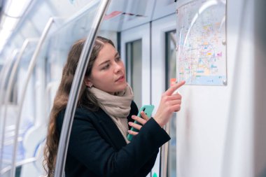 Siyah ceketli ve bej eşarplı odaklanmış genç bir kadın akıllı telefonunu tutarken metro haritasını kontrol ediyor. Modern bir metro treninde rotasını çiziyor..