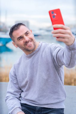 Gri kazaklı sıradan bir adam, marinanın yakınında kırmızı bir akıllı telefonla selfie çekiyor. Poz verirken barış işareti yapıyor. Arka planda tekne ve rıhtım manzarası var. Modern yaşam tarzı 
