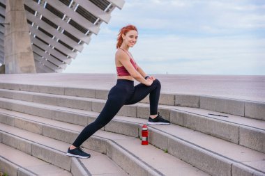 Spor kıyafetleri giyen atletik kızıl saçlı kadın modern bir şehir ortamında bacağını açık merdivenlerde esnetiyor. Parlak mavi gökyüzünün altında egzersiz yapmaya hazırlanan bir su şişesi yakınlarda oturur..