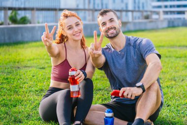 Gülümseyen genç adam ve kadın çimlerin üzerinde oturuyorlar, barış işareti gösteriyorlar ve egzersiz yaptıktan sonra neşeli görünüyorlar. Spor kıyafetleri giyiyorlar ve güneşli bir parkta temiz havanın tadını çıkarıyorlar..
