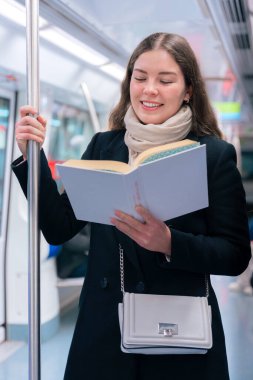 Modern bir metro treninde duran siyah paltolu ve bej eşarplı odaklanmış genç bir kadın metal bir direğe tutunarak kitap okuyor. Seyahat sırasında sakin bir anın tadını çıkarıyor..