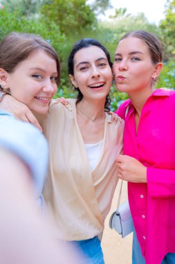 Üç gülen kadın yemyeşil bir parkta selfie çektirmek için poz veriyor. Birbirlerine sarılırlar, mutluluğu ve dostluğu ifade ederler. Canlı renkler ve doğal arka plan neşeli bir atmosfer yaratır.. 