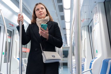 Siyah paltolu ve bej eşarplı genç bir kadın modern bir metro treninin içinde elinde akıllı bir telefonla metal bir direği kavrıyor. Şehirde işe gidip gelirken düşünceli görünüyor.. 