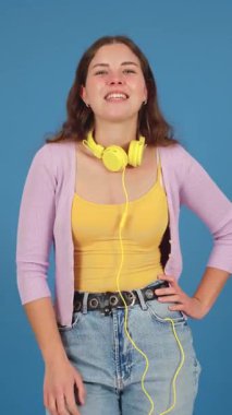  Sarı kulaklık takan ve günlük kıyafetler giyen neşeli genç bir kadın sanki birine hitap ediyormuş gibi doğrudan öne bakıyor. Kendine güveni ve ilgi çekici ifadesi mavi arka planda göze çarpıyor.. 