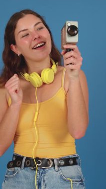 Sarı kulaklık takan ve kolsuz tişörtü olan neşeli genç bir kadın elinde heyecanla klasik bir kamera tutuyor. Mavi arka plan onun neşeli ve nostaljik ifadesini arttırır.. 