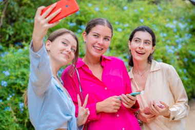 Üç gülen kadın yemyeşil bir parkta selfie çektirmek için poz veriyor. Birbirlerine sarılırlar, mutluluğu ve dostluğu ifade ederler. Canlı renkler ve doğal arka plan neşeli bir atmosfer yaratır.. 