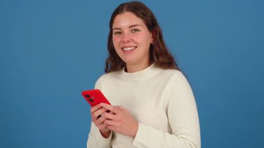 Beyaz bir kazak giyen, kırmızı bir akıllı telefon tutan ve neşeyle gülümseyen neşeli genç bir kadın. Mavi arka plan onun parlak ve çekici ifadesini arttırır..