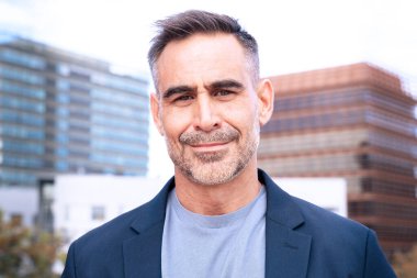 Modern ofis binalarının arka planına güvenle gülümseyen şık takım elbiseli orta yaşlı bir işadamı. Profesyonel ve cana yakın tavırları başarıyı ve liderliği yansıtıyor.. 