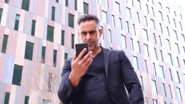 Siyah takım elbiseli kendine güvenen bir işadamı şehir ortamında dikilirken akıllı telefonunu işaret eder. Onun ciddi ifadesi teknoloji, iletişim ve iş iletir..