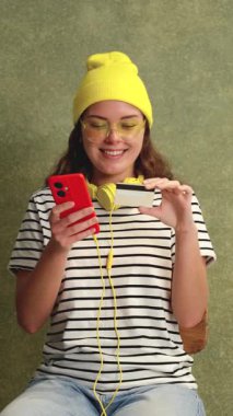 Sarı bereli, güneş gözlüklü, elinde akıllı telefon ve kredi kartıyla gülümseyen son moda genç bir kadın. Sahne çevrimiçi alışveriş ve dijital işlemleri vurguluyor. 