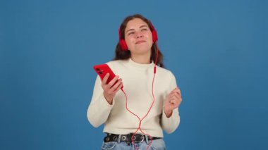 Kırmızı kulaklık takan neşeli genç bir kadın, akıllı telefonuyla müzik dinlerken dans ediyor. O, anlamlı ve enerjik bir jestle ritmi sever. Mavi arkaplan. 