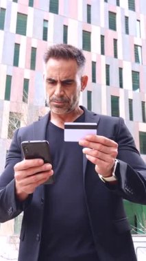 Siyah takım elbiseli profesyonel bir işadamı şehir ortamında akıllı telefonunu kullanırken bir kredi kartı tutuyor. Odaklanmış ifadesi online bankacılık ya da dijital ödeme öneriyor. 