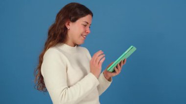 Beyaz kazaklı, yeşil tabletle mavi arka planda etkileşime giren mutlu genç bir kadın. Modern teknolojiyi kullanarak keyif ve odaklanmayı ifade etmek. 