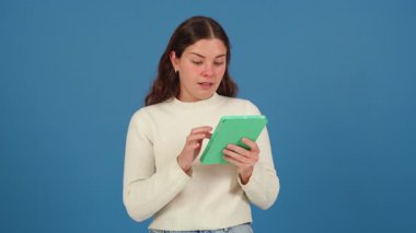 Beyaz kazaklı, yeşil tabletle mavi arka planda etkileşime giren mutlu genç bir kadın. Modern teknolojiyi kullanarak keyif ve odaklanmayı ifade etmek. 