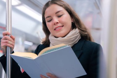 Modern bir metro treninde duran siyah paltolu ve bej eşarplı odaklanmış genç bir kadın metal bir direğe tutunarak kitap okuyor. Seyahat sırasında sakin bir anın tadını çıkarıyor..