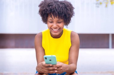 Kıvırcık siyah saçlı ve sarı bluzlu neşeli genç bir kadın açık havada oturmuş, akıllı telefonuna bakarken gülümsüyor. Ağaçların ve binaların bulanık şehir arkaplanı modern bir dokunuş katıyor..