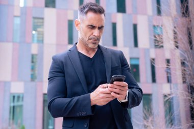 Mavi tişörtlü, gülümseyen orta yaşlı bir işadamı modern bir şehir ortamında akıllı telefon kullanıyor..