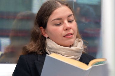  Siyah paltolu ve bej eşarplı genç bir kadın modern bir metro treninde oturmuş, odaklanmış bir kitap okuyor. Şehirdeki seyahati sırasında sakin bir anın tadını çıkarıyor..