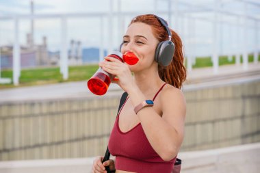 Spor giyim ve kulaklık takan kızıl saçlı atletik bir kadın egzersiz yaptıktan sonra su içiyor. Spor çantası taşıyor ve bulutlu bir gökyüzünün altında modern bir şehir ortamında duruyor.. 