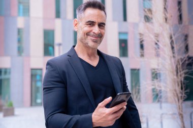 Mavi tişörtlü, gülümseyen orta yaşlı bir işadamı modern bir şehir ortamında akıllı telefon kullanıyor..
