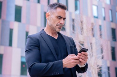 Mavi tişörtlü, gülümseyen orta yaşlı bir işadamı modern bir şehir ortamında akıllı telefon kullanıyor..