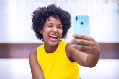 Akıllı telefonuyla selfie çeken genç bir kadın. Modern şehir ortamında binalar ve ağaçlarla süslenmiş şık beyaz bir kıyafet giyerek neşeli bir pozla gülümsüyor..