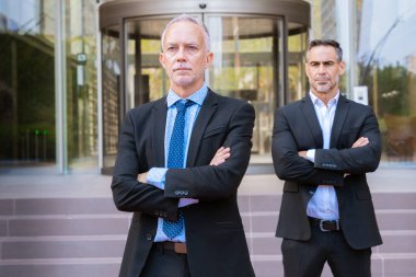 Takım elbiseli iki işadamı modern bir ofis binasının önünde kollarını kavuşturarak güvenle duruyorlar. Profesyonel bir ortamda liderlik, takım çalışması ve kurumsal başarıyı temsil eder..