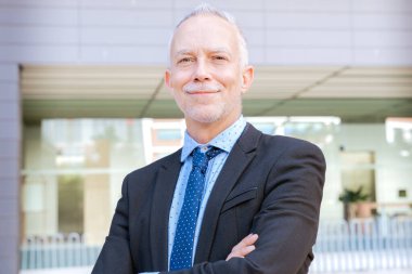 Takım elbiseli profesyonel bir işadamı kollarını kavuşturup modern bir ofis binasının önünde kendinden emin bir şekilde gülümser..