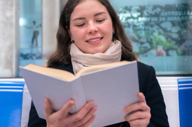  Siyah paltolu ve bej eşarplı genç bir kadın modern bir metro treninde oturmuş, odaklanmış bir kitap okuyor. Şehirdeki seyahati sırasında sakin bir anın tadını çıkarıyor..