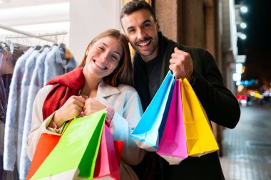 Renkli alışveriş torbaları tutan çift alışverişin tadını çıkarırken gülümsüyor. Mevsim için sıkı giyinmiş bir şekilde etrafları festival ışıklarıyla çevrili, alışveriş çılgınlığının heyecanını gösteriyorlar.