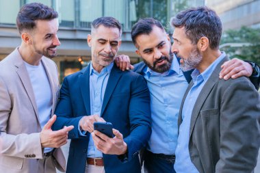 Dört iş adamından oluşan bir grup açık havada toplandı, gülümsüyor ve akıllı telefondan içerikleriyle ilgileniyorlardı. Modern bir şehir ortamında profesyonel ve dostane bir an..