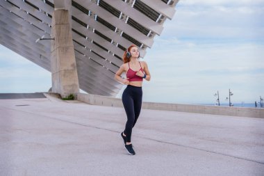 Sahil kenarında koşan kızıl saçlı, zinde bir genç kadın. Spor kıyafetleri giyiyor ve mutlu ve enerjik görünüyor. Arka planda parlak bir gökyüzü ve modern bir şehir ortamı yer alıyor.. 
