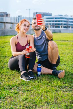 Gülümseyen genç adam ve kadın çimenlerde oturup egzersiz yaptıktan sonra selfie çekiyorlar. Spor kıyafetleri giyiyorlar ve ellerinde su şişeleri var. Şehir parkında rahat bir anın tadını çıkarıyorlar..